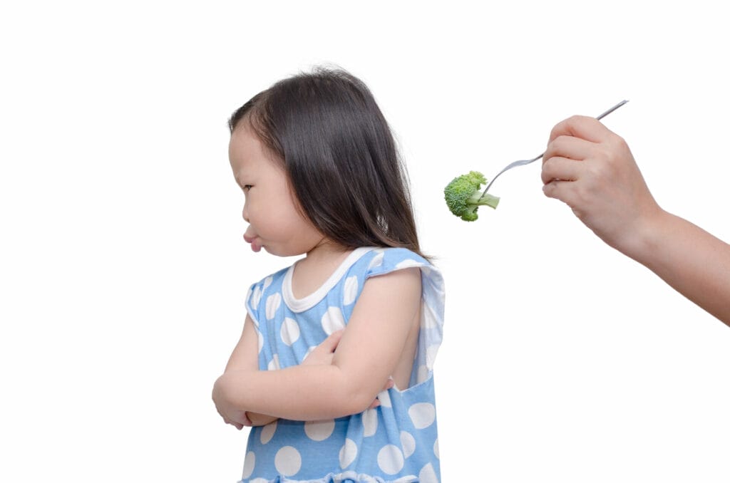 Young girl turning away from a piece of broccoli being handed to her on a fork.